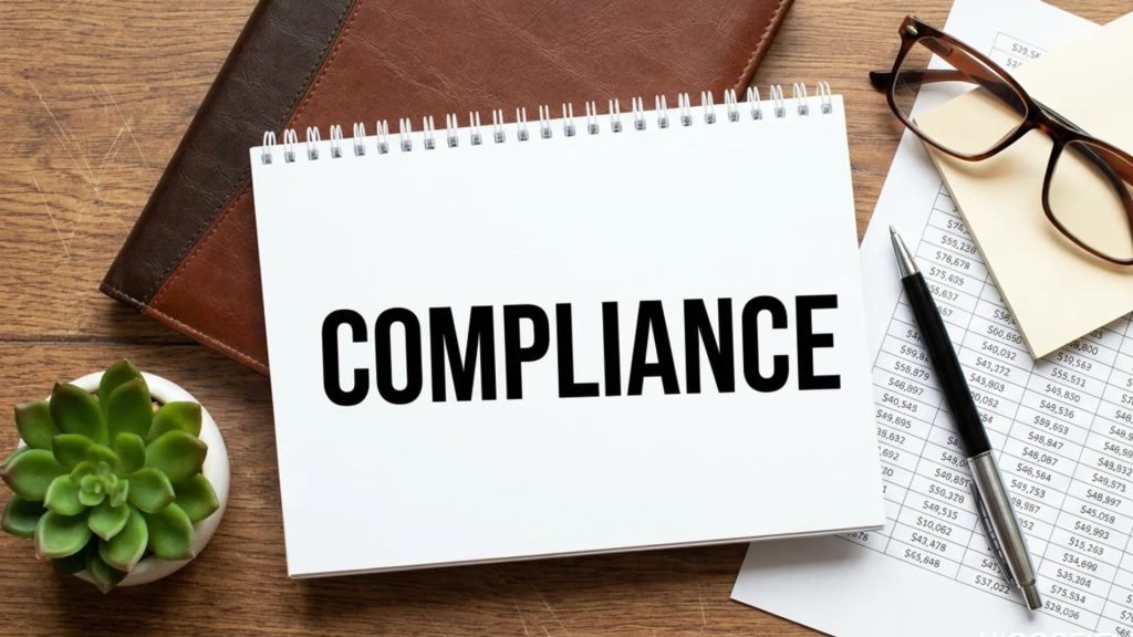 Notebook displaying the word "COMPLIANCE" surrounded by office supplies.