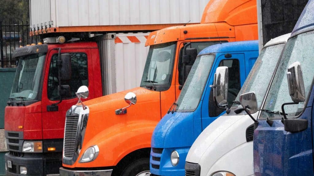 A row of colorful semi-truck cabs, including orange, blue, and white trucks, parked side by side outdoors.